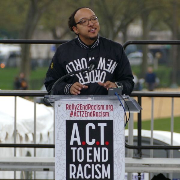a man in a jacket addresses a rally behind a podium reading "ACT to end racism"