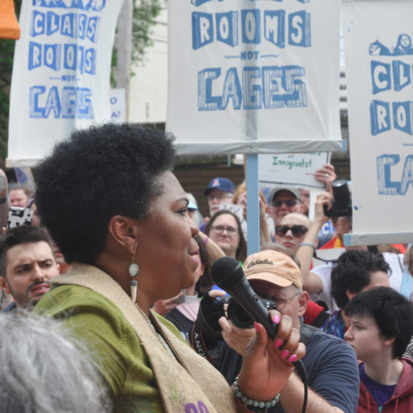 a woman holding a microphone addresses a crowd holding signs that say, "Class rooms not cages"